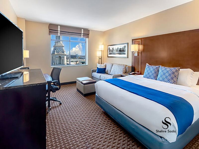 Elegant hotel room featuring a large bed, sitting area with sofas, and panoramic city view through wide windows
